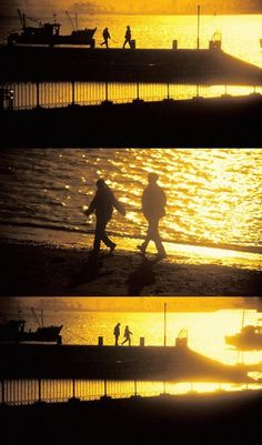 people walking on the beach at sunset with boats in the water and one man carrying a surfboard