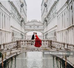 a man and woman in red dress standing on bridge over water with buildings behind them