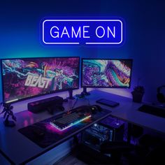 two computer monitors sitting on top of a desk in front of a neon sign that says game on