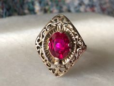 This stunning vintage ring is a beautiful example of antique jewelry craftsmanship, made of 10k gold and featuring a captivating, vibrant red faceted stone at its center. The stone, rich and intense in color, is securely set within an ornate setting that reflects a blend of late Victorian to Art Deco style, characterized by intricate lacy motifs and a delicate filigree pattern. The combination of the deep red stone and the intricate goldwork creates a striking visual contrast, enhancing the over Formal Red Ruby Ring Stamped 14k, Red Victorian Filigree Ring For Anniversary, Victorian Red Oval Filigree Ring, Collectible Red Ruby Ring In 14k Gold, Ornate Gold Ruby Ring, Antique Oval Red Ruby Ring, Red Filigree 14k Gold Jewelry, Red 14k Gold Filigree Jewelry, Antique 14k Gold Ruby Ring With Birthstone