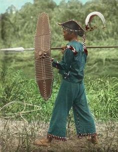 a young boy dressed in native american garb holding a fan and arrow while walking through the grass