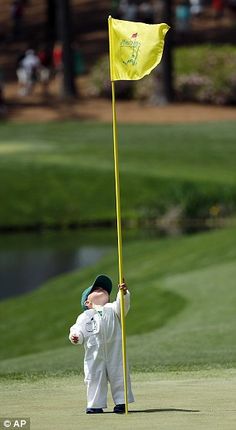 a young boy holding a yellow flag on top of a green golf course in the sun