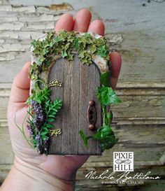 a hand holding a miniature house made out of wood and plants on the outside, in front of a brick wall