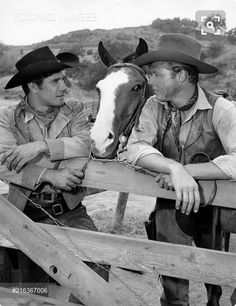 two men in cowboy hats leaning on a fence next to a horse