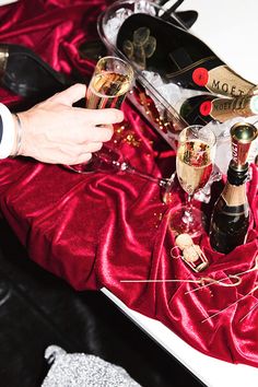 a man is holding two glasses of champagne while sitting at a table with other wine bottles