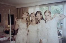 three girls in white dresses standing next to each other near a bed with a teddy bear on it