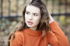 a woman in an orange sweater is holding her hair up and looking at the camera