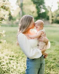 a woman holding a baby in her arms and kissing it's face while standing in the grass