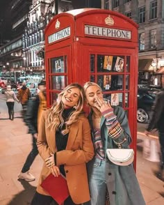 two women standing next to each other in front of a red phone booth on the street