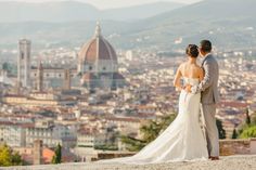 a bride and groom standing on top of a hill looking at the city in the distance