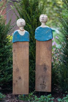 two wooden statues sitting next to each other in front of bushes and trees, one with a woman's face painted on it