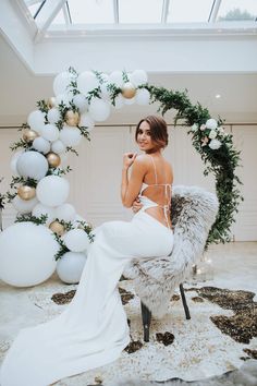 a woman sitting on a chair in front of a balloon arch with white and gold decorations