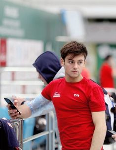 a man in a red shirt standing next to a metal barricade and looking at his cell phone