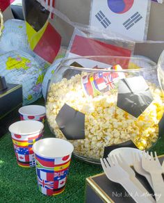 a bowl filled with popcorn next to plastic cups and utensils on top of grass