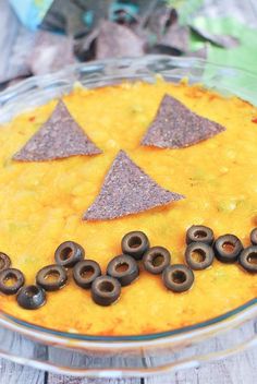 a bowl filled with dip and black olives on top of a wooden table next to chips