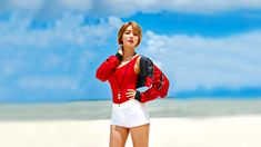 a woman in red shirt and white shorts standing on beach with headphones to her ear