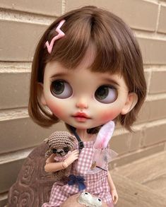 a close up of a doll holding a stuffed animal near a brick wall with a building in the background