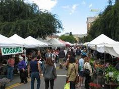 many people are walking through an outdoor market with white tents and trees in the background