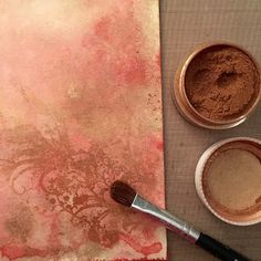 two bowls with powder and a brush next to it on a piece of paper that has been painted