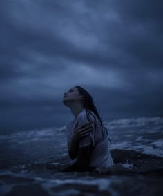 a woman sitting in the water with her eyes closed, looking up at an overcast sky