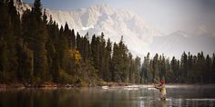 a man standing in the middle of a lake holding a fly fishing rod with mountains in the background