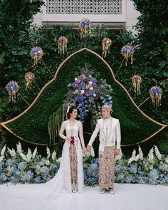 two people standing next to each other in front of a flower covered area with blue and white flowers