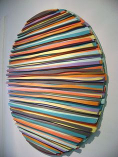 a circular sculpture made out of strips of colored paper on a white wall in an art gallery