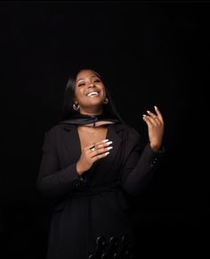 a woman standing in front of a black background with her hands together and smiling at the camera