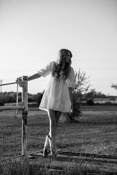 black and white photograph of woman leaning on fence post in grassy field with trees behind her