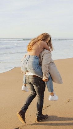 a woman carrying a child on her back while walking along the sand at the beach