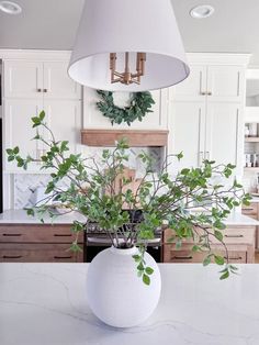 a white vase filled with green plants on top of a counter next to a lamp