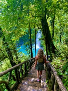 a woman walking up some stairs in the woods with trees on either side and blue water running down it