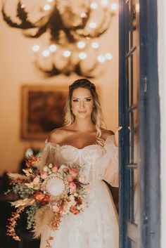 a woman standing in front of a window holding a bridal bouquet and looking at the camera