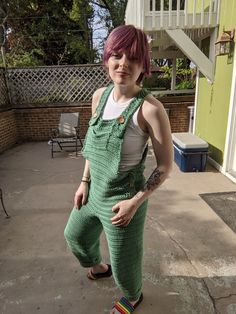 a woman with pink hair and green overalls posing for the camera in front of a house