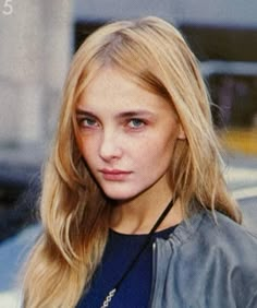 a woman with long blonde hair wearing a leather jacket and looking at the camera while standing in front of a car