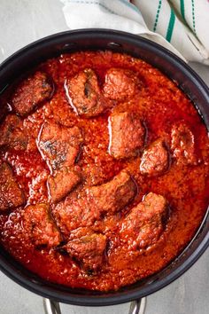 a pan filled with meat and sauce on top of a white cloth next to a fork