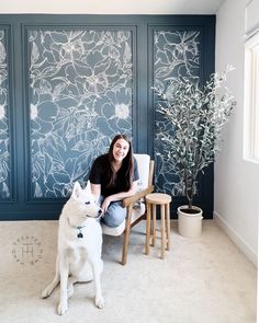 a woman sitting in a chair next to a white dog and wall with flowers on it