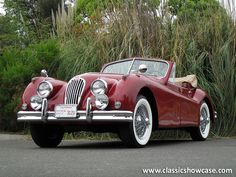 an old red sports car parked in front of some tall grass and bushes with the top down