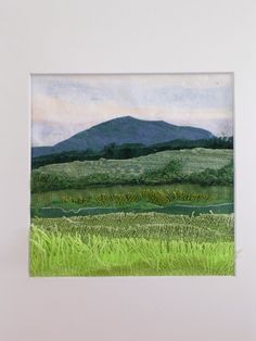 a painting hanging on the wall in front of a white frame with green grass and mountains