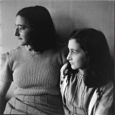 two women sitting next to each other in front of a wall and one is wearing glasses