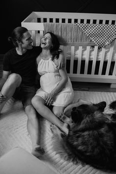 a man and woman sitting next to a dog on a bed with a crib in the background