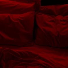 a bed with red sheets and pillows on top of it in a room that is dark