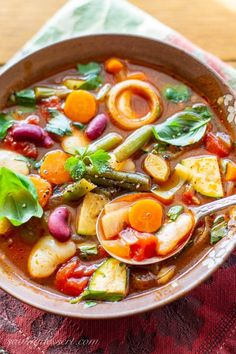 a bowl filled with vegetable soup on top of a red and green table cloth next to a wooden spoon