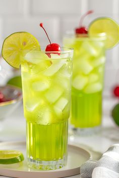 two glasses filled with limeade and cherries sitting on a white plate next to lemon slices