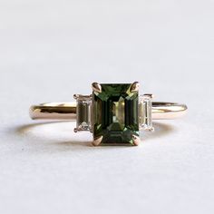 Emerald cut green tourmaline adorned with baguette diamonds Metal: 14K rose gold Stone: Green Tourmaline Stone Size: 8 x6 mm Stone Weight: 1.7 Carat Accent Stone: Baguette Diamonds: Diamond Size: 4.5 x 2 mm (2) Diamond Color and Clarity: GHI, SIDiamond weight: 0.12 carat each Total Diamond Weight: 0.25 carat FREE SHIPP Emerald Gemstone Engagement Ring Gold, Tourmaline And Diamond Ring, Green Engagement Ring And Wedding Band, Wedding Ring With Emerald Accents, Engagement Rings Emerald Cut Three Stone, Vintage Green Wedding Ring, Green Gem Rings, Green Emerald Cut Engagement Ring, Emerald Cut Gemstone Ring