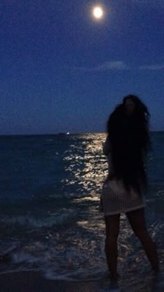 a woman standing on top of a beach next to the ocean under a full moon