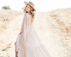 a woman in a white dress and hat is standing on a dirt road with her back to the camera