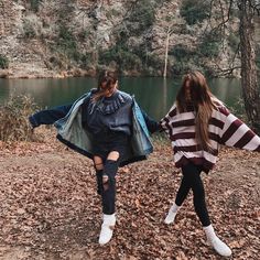 two young women walking in the woods next to a body of water with trees behind them