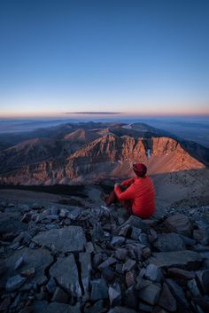 a man sitting on top of a rocky hill next to a mountain range at sunset