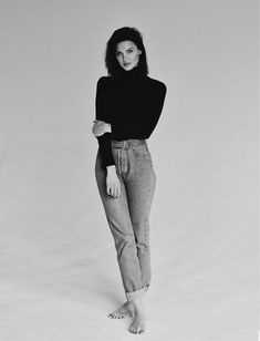 a woman in black shirt and jeans standing with her hands behind her back, looking at the camera